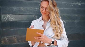 en kvinna i glasögon lutande på en sten vägg är innehav en notbook och skrivning på den video