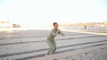 Fitness woman in sports wear doing lunges on step, and stretching legs on summer park near seaside. video