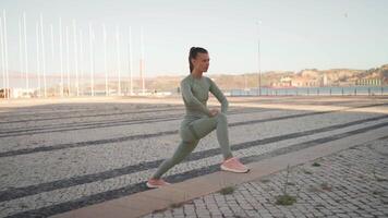 Fitness woman in sports wear doing lunges on step, and stretching legs on summer park near seaside. video