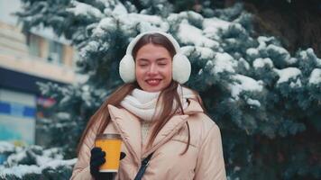 Close up portrait young woman with fur earmuffs standing with coffee cup on spruce tree background. winter day outdoor. video