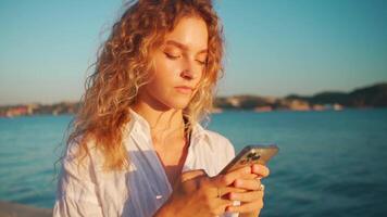 femme porter blanc chemise avec frisé cheveux en utilisant téléphone intelligent app près rivière. video
