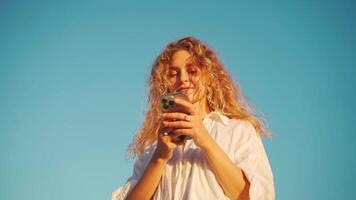 mulher vestem branco camisa com encaracolado cabelo usando Smartphone aplicativo perto rio. video