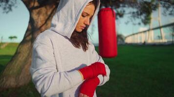 Woman shadow boxing with her hands wrapped in red boxing tapes in park with punching box. video