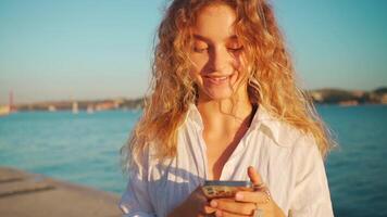 Woman wear white shirt with curly hair using smartphone app near river. video