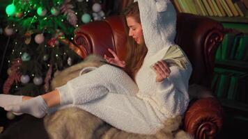 Young woman headphones sits leather armchair near christmas tree holds smartphone in hand chooses a song. video