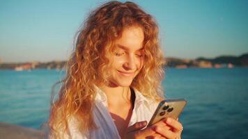 mujer vestir blanco camisa con Rizado pelo utilizando teléfono inteligente aplicación cerca río. video