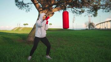 Woman shadow boxing with her hands wrapped in red boxing tapes in park with punching box. video