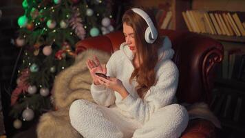 Young woman headphones sits leather armchair near Christmas tree holds smartphone in hand chooses a song. video