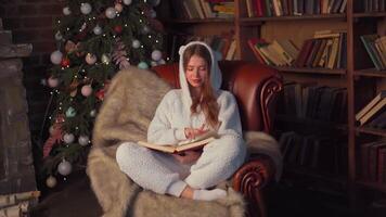 mujer leyendo libro sentado acogedor cuero Sillón con Navidad árbol en antecedentes. video