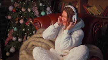 Young woman headphones sits leather armchair near Christmas tree video