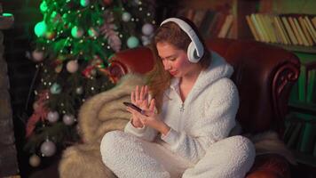 Young woman headphones sits leather armchair near Christmas tree holds smartphone in hand chooses a song. video