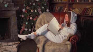 Woman reading book sitting cozy leather armchair with christmas tree in background. video