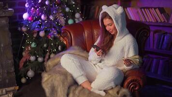 Young woman headphones sits leather armchair near Christmas tree holds smartphone in hand chooses a song. video