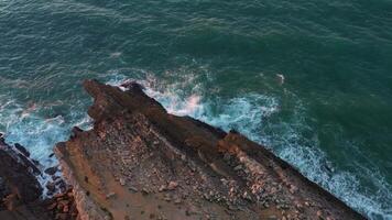 aéreo zumbido ver de escabroso rocoso línea costera con olas estrellarse y rotura en contra rocas a amanecer. video