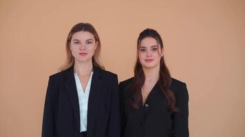 Medium portrait shoot of two young business woman dressed black jacket standing studio over pastel color background. She is shy laughs talk and speak. Female modern business person video