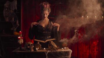 Halloween witch with cauldron and magic book. Beautiful woman flowers in hair conjuring, making witchcraft. Standing spooky dark room. Enchantress prepare love potion Slow motion. Handheld video