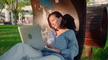 Happy brunette woman using laptop computer while sitting in summer park. Young female freelancer working outdoors in city park. Caucasian businesswoman at remote work. Business person. Freelancer video