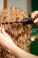 Hands of hairstylist curl wavy hair of young woman using a curling iron for hair curls in the beauty salon rear view. photo