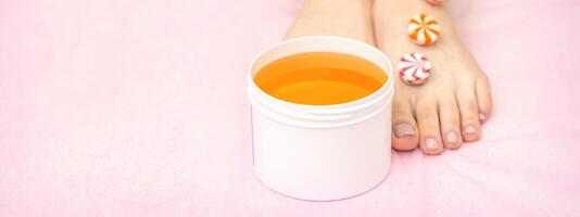 Round candies lying in a row on female feet with white jar with sugar paste on pink background with copy space, depilation concept. photo