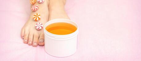 Round candies lying in a row on female feet with white jar with sugar paste on pink background with copy space, depilation concept. photo