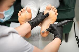 Peeling female feet. Hands of podiatry specialist in black gloves using special grinding equipment polish female foot from callus and corn in the beauty salon. photo