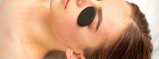 Black massage stones lying on the eyes of the young caucasian woman. Facial massage in a spa. photo