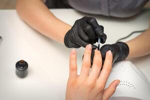 Painting nails of a woman. Hands of Manicurist in black gloves applying transparent nail polish on female Nails in a beauty salon. photo