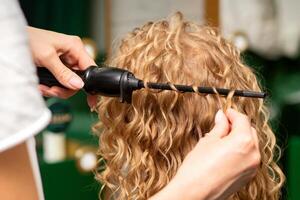 Hands of hairstylist curl wavy hair of young woman using a curling iron for hair curls in the beauty salon rear view. photo