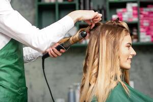 creando chinos con curling hierros. peluquero hace un peinado para un joven mujer con largo rojo pelo en un belleza salón. foto