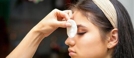 Side view of a makeup artist covers female eye with a cotton pad using airbrush making makeup foundation on her face in a beauty salon. photo