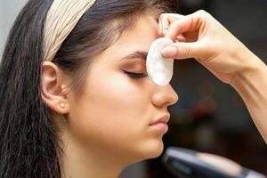 Side view of a makeup artist covers female eye with a cotton pad using airbrush making makeup foundation on her face in a beauty salon. photo