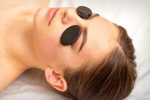 Black massage stones lying on the eyes of the young caucasian woman. Facial massage in a spa. photo