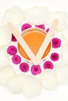 Jar of wax with wooden sticks, pink flowers, and white petals on white background. Concept of depilation. Flat lay top view. photo