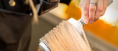 peluquero mano el secado rubio pelo con un secador de pelo y redondo cepillo en un belleza salón. foto