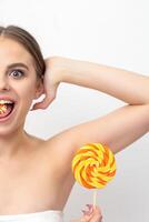 Happy expression young nude woman with candy in mouth holds lollipop near armpit on white background, epilation, depilation, hair removal concept. photo