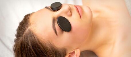 Black massage stones lying on the eyes of the young caucasian woman. Facial massage in a spa. photo