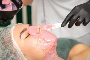 el cosmetóloga aplicando un alginato máscara a el cara de un joven mujer en un belleza salón. foto