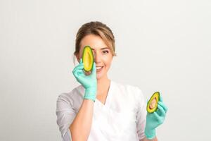 Portrait of young female nutritionist doctor with beautiful smile posing at camera hiding eye behind half avocado on white background, copy space. Benefits of proper nutrition. photo