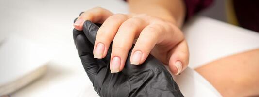 examen de cuidado uñas manos de manicura Maestro en negro guantes examinando hembra transparente uñas en manicura salón. foto