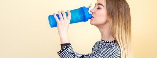 Thirsty woman with blue plastic bottle against a light background. A beautiful young caucasian businesswoman in a blouse drinking water on yellow. photo