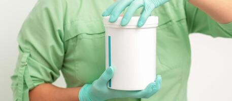 Hands in protective gloves of beautician open a white body cream jar on white background. photo