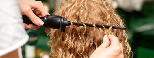 Hands of hairstylist curl wavy hair of young woman using a curling iron for hair curls in the beauty salon rear view. photo