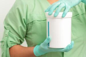 Hands in protective gloves of beautician open a white body cream jar on white background. photo