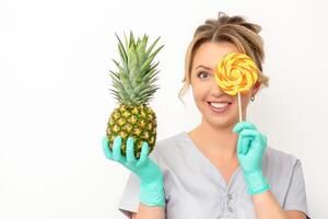 Beautiful smiling beautician holding fresh pineapple and cover her eye with lollipop over white background. Skincare cleansing eco organic. photo