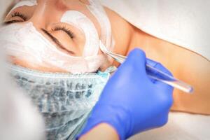 cara peladura a el cosmetólogo. facial tratamiento. el cosmetólogo aplica un limpieza cara máscara a el hembra paciente. foto