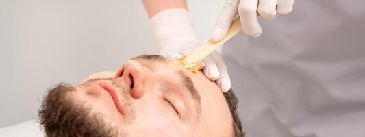 cosmetólogo aplicando cera pegar Entre Cejas durante el procedimiento de depilación en el belleza salón. foto