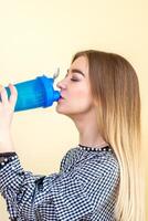 Thirsty woman with blue plastic bottle against a light background. A beautiful young caucasian businesswoman in a blouse drinking water on yellow. photo