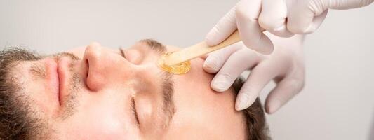 cosmetólogo aplicando cera pegar Entre Cejas durante el procedimiento de depilación en el belleza salón. foto