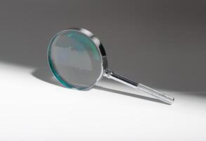 Silver magnifying glass on glass background. Shadow and light play on circular lens. Concept of photo