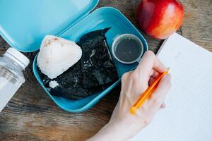 Asian student's lunchbox made of onigiri, apple and soy sauce with a bottle of water, On a wooden background. A student writes down his plans in a notebook during a break. photo